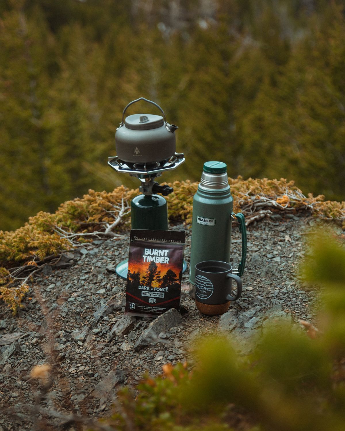 A kettle on a portable camping stove beside a green flask, coffee bag and mug on a rocky forest surface.