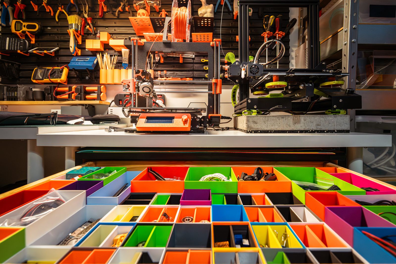 A desktop 3D printer surrounded by tools, filament and colorful storage bins in a workshop.